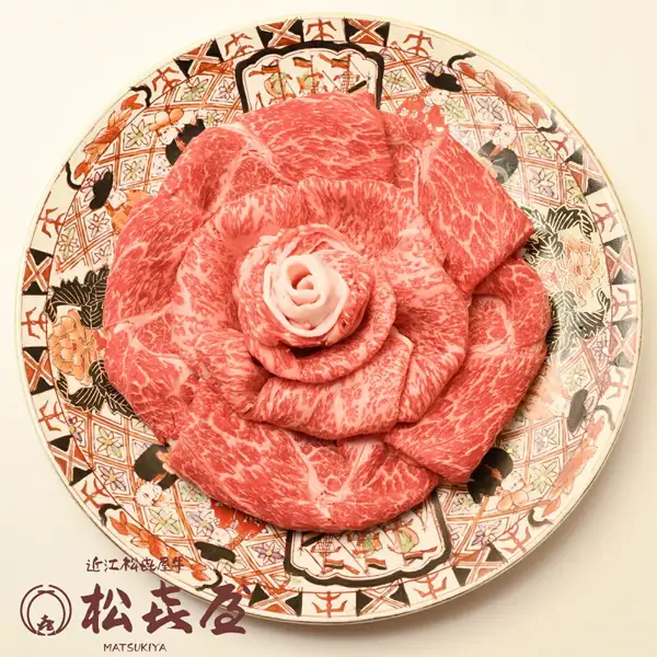 Product photo of Omi beef sukiyaki slices (loin, momo and bara) arranged in the shape of a rose on a round plate with a red, orange and black floral pattern. The store logo with the text “近江松喜屋牛” and “松喜屋 MATSUKIYA” appears in the image (shown by the store). Tokusen Omi Gyu Sukiyaki Beef, Loin, Momo & Bara, 1kg