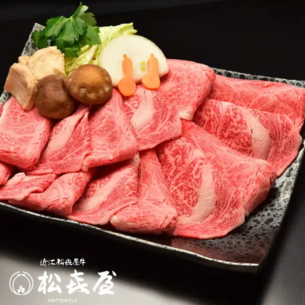 【特選】近江牛 すき焼き用 ロース・モモ・バラ1kgのスライス肉を黒い角皿に盛り付けた写真。肉の上と奥に白菜・三つ葉・玉ねぎ・しいたけ・にんじんなどの野菜・きのこ類（ほか肉以外の食材）を添えているが、これらは商品に含まれない。画像内に店舗ロゴ「近江松喜屋牛」「松喜屋 MATSUKIYA」の文字あり（店舗が画像に入れた表示）。