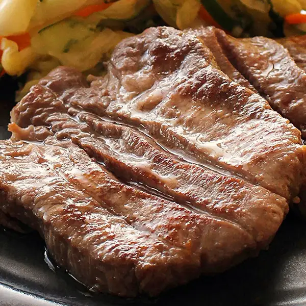 Close-up photo of several thick slices of grilled beef tongue on a black plate, with a vegetable side dish that looks like cucumber, cabbage and carrot behind them. There is no text in the image. 肉のいとう Aged Thick-Cut Beef Tongue, Salt-Seasoned, 500g