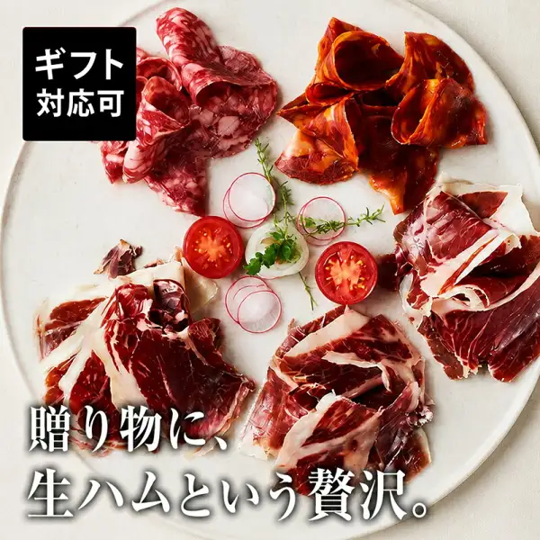 イベリコ豚 生ハム（生火腿）5种食べ比べセット（イベリコ屋）的商品照片。白色圆盘上摆着切片的萨拉米、辣香肠和3种生火腿，配有小萝卜、番茄、洋葱和香草。店铺在图片中加入的标示「ギフト対応可」「贈り物に、生ハムという贅沢。」。