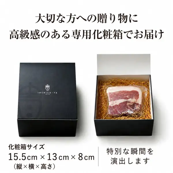 生ハム専用豚肉 アンダルシアポーク ミニ原木の化粧箱の商品写真。左に店舗ロゴ「IBERICO-YA」入りの黒い化粧箱、右に蓋を開けた箱があり、わらのような緩衝材の上に透明な袋に入った生ハムのブロックが収まっている。店舗が画像に入れた表示「大切な方への贈り物に」「高級感のある専用化粧箱でお届け」「化粧箱サイズ 15.5cm×13cm×8cm（縦×横×高さ）」「特別な瞬間を演出します」。
