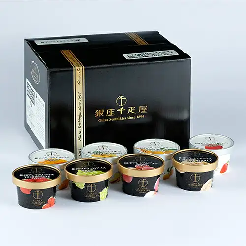 黑色盒子前方，前排摆放4个金色盖子的黑色杯装（冰淇淋）、后排摆放4个透明盖子的白色杯装（雪葩）的商品照片。前排盖子上有「銀座プレミアムアイス」字样、「TOCHIOTOME」「MUSCAT」「PEACH」「CUSTARD VANILLA」英文及水果等图案，后排盖子上有芒果、菠萝、柚子与蜂蜜、草莓的图案。盒子和杯子上印有店铺标志「銀座千疋屋」「Ginza Sembikiya since 1894」（店铺加入图片的标示）。銀座プレミアムアイス＆ソルベ（冰淇淋与雪葩） 8個入（パティスリー銀座千疋屋）