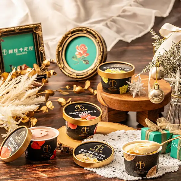 Photo of 4 ice cream flavor cups set on a wooden table with Christmas decorations and picture frames. It shows Tochiotome (pale pink) and Custard Vanilla (cream-colored, being scooped with a gold spoon) with their lids off, Peach with its lid on, and Muscat on a small stand. The lids read “銀座プレミアムアイス”, “TOCHIOTOME”, “PEACH” and “CUSTARD VANILLA”, and the cups and a frame carry the shop's logo “銀座千疋屋”, “GINZA SEMBIKIYA” and “Since 1894” (a mark the shop placed in the image). Ginza Premium Ice & Sorbet, 8 Pieces (パティスリー銀座千疋屋)