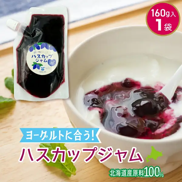 Serving-example photo of Additive-Free Haskap Jam, Hokkaido, 160g (Tokachi Berry Farm). On a wooden table, dark purple haskap jam is spooned over yogurt in a white bowl with a white spoon, with one stand-up pouch with a white cap at the back left and mint leaves in front and on the left (the yogurt and mint leaves are not included in the product). Text the shop placed in the image includes “160g入 1袋” (160g, 1 pouch), “ヨーグルトに合う！” (goes well with yogurt), “ハスカップジャム” (haskap jam) and “北海道産原料100%” (100% Hokkaido-grown ingredients), with a mark in the shape of Hokkaido.