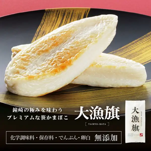 黒い漆器の皿に金色の筆跡模様を背景に、焼き色のついた大漁旗の笹かまぼこ2枚を重ねた写真。右下に大漁旗の袋入り商品も写る(鐘崎 笹かまぼこ 大漁旗特選詰め合せ TF-3A7 13枚入)。店舗が画像に入れた表示「鐘崎の極みを味わう プレミアムな笹かまぼこ」「大漁旗」「化学調味料・保存料・でんぷん・卵白 無添加」
