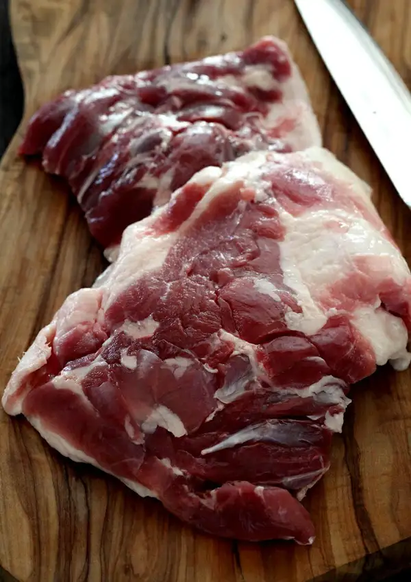 Photo of two pieces of raw duck thigh meat on a wooden cutting board, showing red meat and white fat, with the tip of a knife at the upper right (thigh meat of the 豊代 鴨三昧 岩手がも鍋セット)