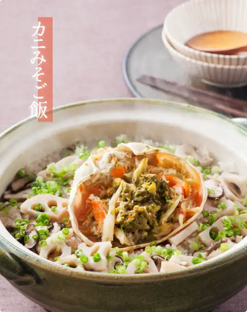 土鍋で炊いたカニみそご飯の写真（盛り付け例）。ご飯の上に、蟹味噌と蟹の身をのせた甲羅、れんこん、きのこ、刻みねぎがのっている。手前に小さな器と箸。店舗が画像に入れた表示「カニみそご飯」