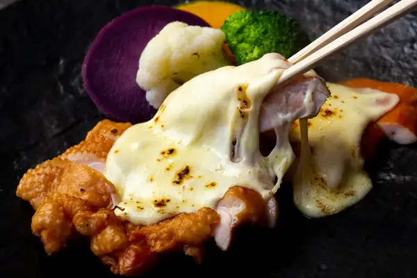 Serving-example photo of smoked chicken topped with melted cheese on a black slate-style plate, lifted with chopsticks (Pure-Bred Nagoya Cochin Smoked 4-Kind Set). Purple sweet potato, cauliflower, broccoli and pumpkin sit behind it. The cheese and vegetables are not included in the product.