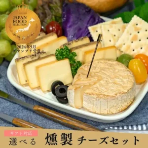 皿に並べた燻製チーズ（煙神 選べる燻製チーズ詰め合わせ）の商品写真。ブラックペッパーをのせた長方形のチーズ、燻製チーズ、切り分けたカマンベールタイプのチーズが写っている。皿にはクラッカー・ぶどう・オリーブ・ミニトマト・パセリ・紫キャベツも盛られているが、これらは商品に含まれない盛り付け例。店舗が画像に入れた表示：「JAPAN FOOD SELECTION」「2024年5月 グランプリ受賞」「ギフト対応」「選べる 燻製チーズセット」