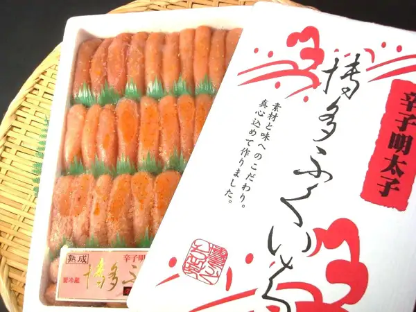 Product photo of 築地丸中 特上博多ふくいち粒々辛子明太子1kg (Tsukijimarunaka Tokujo Hakata Fukuichi Tsubutsubu Karashi Mentaiko 1kg): orange-tinted pink spicy mentaiko roe sacs in a white tray, laid out in three rows separated by green dividers, with the package lid laid over the right side of the tray. The lid is printed with “辛子明太子”, “博多ふくいち” and “素材と味へのこだわり。真心込めて作りました。” as package lettering.