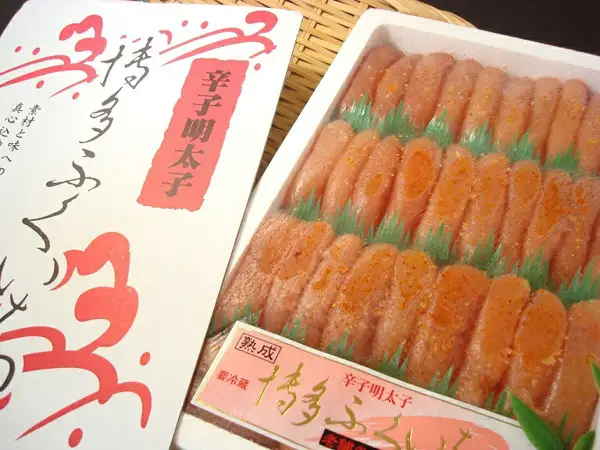 Photo of the contents of 築地丸中 特上博多ふくいち粒々辛子明太子1kg (Tsukijimarunaka Tokujo Hakata Fukuichi Tsubutsubu Karashi Mentaiko 1kg): orange-tinted pink spicy mentaiko roe sacs filling the tray in rows separated by green dividers, with orange grains visible on the surface. A label in front reads “熟成”, “要冷蔵”, “辛子明太子” and “博多ふくいち”, and part of the lid is visible on the left (some of its lettering is cut off).