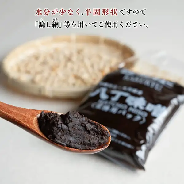Product photo of Kakukyu Hatcho Miso, Commercial Use, 500g × 2 Bags. On a white surface, dark brown, semi-solid Hatcho miso sits on a wooden spoon in front, with one dark commercial-use pack at the back right and a blurred bamboo basket of soybeans at the back left (the soybeans in the basket are not included with the product). The text “八丁味噌” on the bag is visible but blurred. Text the shop placed in the image reads “水分が少なく、半固形状ですので「漉し網」等を用いてご使用ください。” (it has little moisture and is semi-solid, so please use a strainer or similar), with the readings “こし” and “あみ” printed above “漉し網”.