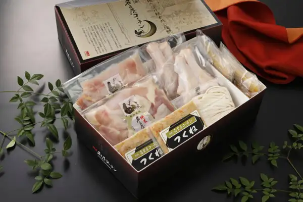 純系名古屋コーチン 旨塩鍋 4〜5人用 うどん付きの商品写真。黒い化粧箱の中に、袋入りのカット肉2パックと骨付きの手羽元、「つくね」と書かれたラベルのパック2つ、うどん、淡い黄色の袋が収まっている。開いたふたには「名古屋コーチン」の文字と鶏の絵の入った紙がある