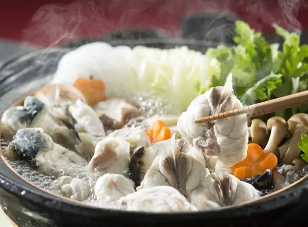 Photo of a steaming black pot in which pieces of fugu, carrots, mushrooms, leafy greens and white vegetables are simmering, with chopsticks lifting a piece of fugu (ふく一 Torafugu Sashimi and Fugu Hot Pot Set, 2–3 Servings). The carrots, mushrooms, leafy greens and white vegetables in the photo are not included in the product