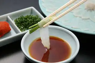 筷子夹着卷着青葱的河豚刺身蘸入盛有褐色酱汁的小碗的照片，左侧有红叶泥和葱花，后方是河豚刺身的盘子（ふく一 とらふぐ刺身・ふぐ鍋セット 2〜3人前）