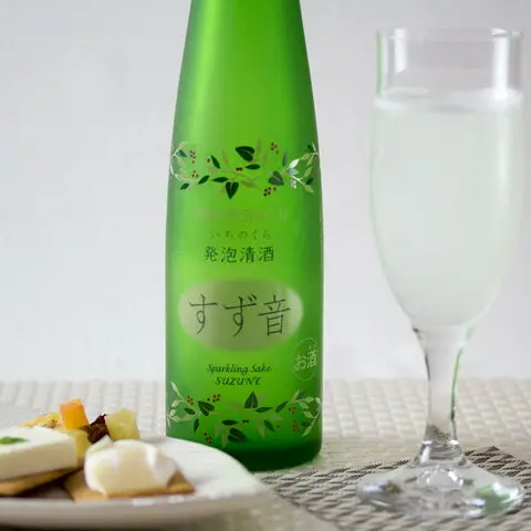 緑色の瓶に葉と赤い実の模様が描かれた一ノ蔵 発泡清酒 すず音 300mlの商品写真。瓶のラベルには「いちのくら」「発泡清酒」「すず音」「Sparkling Sake SUZUNE」「お酒」の文字がある。右には白く濁った酒を注いだ細長いグラス、左下にはチーズや果物をのせたクラッカーの皿が写っている。