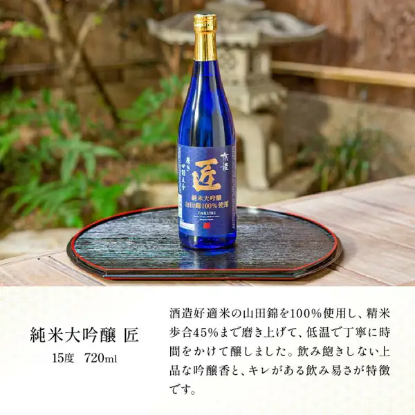 Product photo from the Kyohime Shuzo (京姫酒造) sake “Takumi” (匠) tasting set: one blue bottle of Junmai Daiginjo Takumi standing on a black lacquered tray with a stone garden lantern behind. Text the shop placed in the image: “純米大吟醸 匠” (Junmai Daiginjo Takumi), “15度 720ml” (15 degrees, 720ml), “酒造好適米の山田錦を100%使用し、精米歩合45%まで磨き上げて、低温で丁寧に時間をかけて醸しました。飲み飽きしない上品な吟醸香と、キレがある飲み易さが特徴です。” (Made with 100% Yamada Nishiki, a rice suited to sake brewing, polished to a ratio of 45%, and carefully brewed over time at low temperature. It features an elegant ginjo aroma you won't tire of and a crisp, easy-drinking quality.)