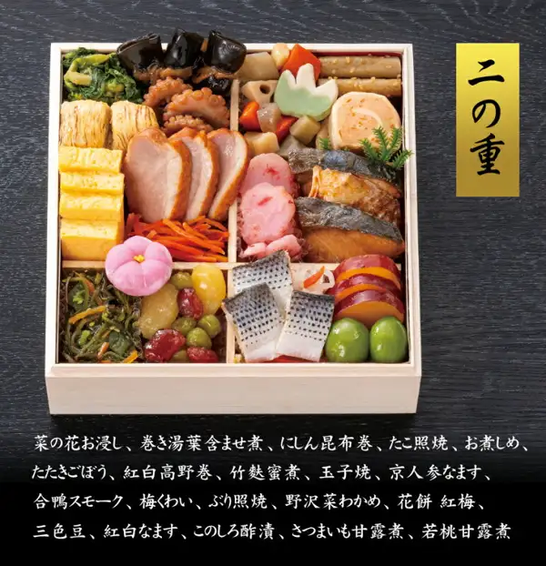 可看清第二层内容的俯拍照片（美食サークル 京都祇おん江口监修 おせち「祝」三段重）：装有厚蛋烧、烟熏鸭肉、照烧鰤鱼、红白萝卜丝和三色豆等。图片中有店铺加入的标示“二の重”，以及以菜の花お浸し、巻き湯葉含ませ煮、にしん昆布巻、たこ照焼开头的20道菜名一览
