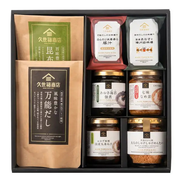 Product photo of the Kuzefuku Shoten Gohan no Otomo Gift (with gift box, 8-item set). Inside a black gift box are two paper pouches of kombu dashi and all-purpose dashi, two miso soup packets (pork miso soup and aosa seaweed miso soup), and four jars (aosa nori tsukudani, shichimi nameko, okazu miso, and shake-shake mentaiko flakes), eight items in total. Text printed on the product packaging includes "久世福商店" and "国産具材使用".