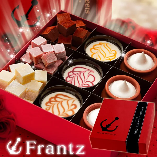 Product photo of Kobe Franz Kobe Minatomachi no Gogo. Inside a red gift box divided into nine compartments are 3 unbaked cheesecakes with orange, red, and caramel-colored patterns, 3 pots of pudding topped with cream, and strawberry, milk, and plain nama chocolates. A red box with a black anchor mark sits in front, with the text "Frantz Sweets Gift" and "www.frantz.jp" on its band and the shop logo "Frantz" at the lower left.