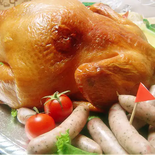 サンライズファーム Roast Smoked Chicken, Whole Bird: Close-up product photo of a whole roast smoked chicken with a golden-brown skin, shown as a serving example. In front are several sausages with visible herb flecks, two cherry tomatoes and a small red flag pick. The cherry tomatoes and flag pick are not included. The type of sausage is a surprise, so the type pictured may differ from what arrives