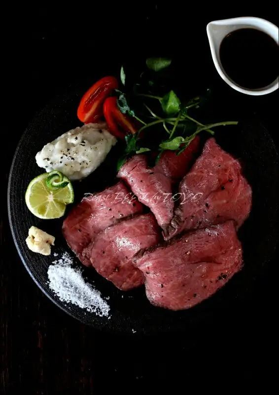 Serving-suggestion photo of sliced roast beef on a black round plate with cherry tomatoes, watercress, a white condiment, a halved lime, salt and a small pitcher of dark sauce (豊代 A4/A5 Kuroge Wagyu Premium Roast Beef, approx. 350g, Tougarashi cut, with special sauce). The image carries the text “Roast BEEF by TOYO”