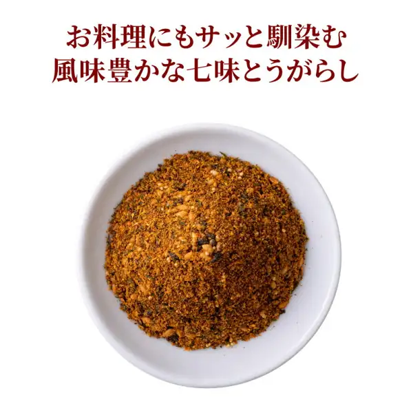 Photo of shichimi togarashi heaped in a small white dish: a red-orange powder mixed with white and black grains and small green flakes (the contents of the shichimi in the 舞妓はんひぃ～ひぃ～ Set). Text the shop placed in the image: “お料理にもサッと馴染む風味豊かな七味とうがらし”