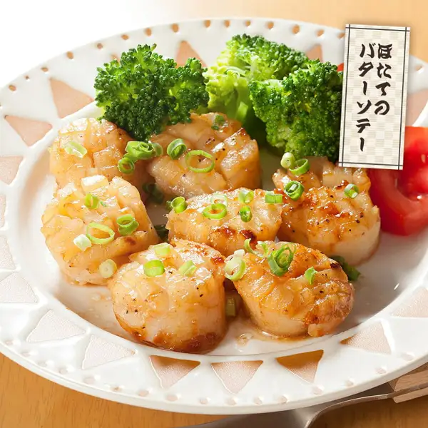 Serving-example photo for Hokkaido Scallop Adductor Muscle Toku-A/A Grade 1kg: butter-sautéed scallop adductor muscles with a browned surface arranged on a round white plate with a scalloped rim, garnished with broccoli, chopped green onions and tomato. A sign at the upper right reads “ほたてのバターソテー” (butter-sautéed scallops).
