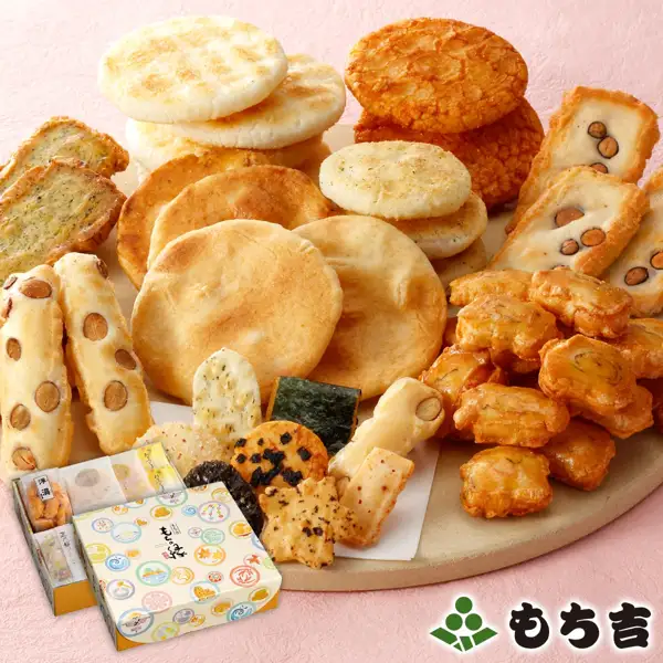 Product photo of the Mochikichi Tasting Set Mochi no En Aji Madoka: round senbei, bean-studded okaki and other rice crackers piled on a large round board against a pink washi-style background, with a patterned outer box in front. Labels added to the image by the shop: the shop logo “もち吉” at the lower right, and “もち吉” and “もちの縁” on the outer box.