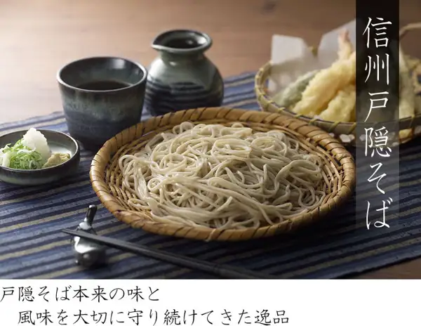 照片：竹箕上盛放的荞麦面，前方有筷子和筷架，后方是蓝色碗中的蘸汁（つゆ）和酒壶，左侧小碟中有葱、萝卜泥等佐料，右后方是盛有天妇罗的篮子。右侧竖写着「信州戸隠そば」，下方白色横条上写着「戸隠そば本来の味と風味を大切に守り続けてきた逸品」（店舗在图片中加入的标示）。蘸汁（つゆ）、佐料和天妇罗是盛放示例，商品页面的套装内容中没有记载。信州戸隠そば お試しセット