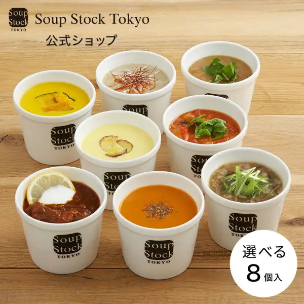 Soup Stock Tokyo 選べる8スープセットの商品写真。木のテーブルの上に、白いカップに入った黄色・薄黄色・オレンジ色・赤茶色・トマト色・茶色など色の異なる8つのスープが並び、ネギ・香草・レモン・赤い糸状の唐辛子などがのっている。カップと具の盛り付けは例で、商品に含まれるのは冷凍スープのパック。店舗が画像に入れた表示：「Soup Stock Tokyo」ロゴ、「公式ショップ」「選べる 8個入」