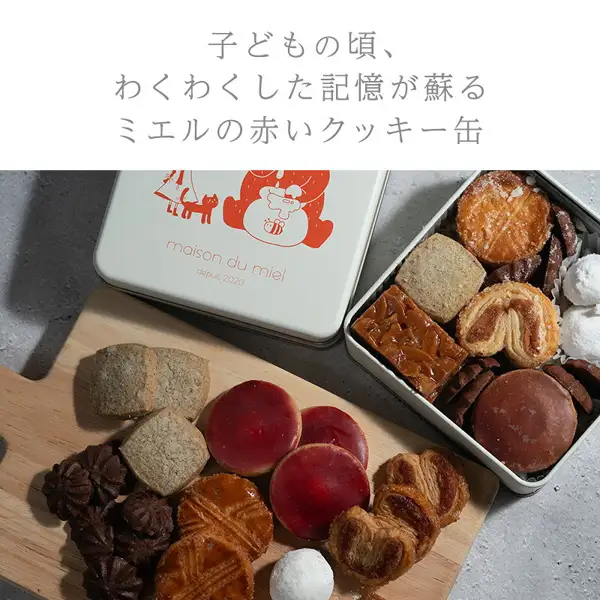 白色罐盖，以及摆放在罐中和砧板上的曲奇的maison du miel ミエルの赤いクッキー缶（红色曲奇罐）内容物商品照片。罐中装有パルミエ和フロランタン・サブレ等，砧板上摆放着赤い実のサブレ、ガレット・ブルトンヌ、パルミエ、ディアマン・アールグレイ、ポッシェ・ショコラ、ブール・ド・ネージュ。店铺在图片中加入的文字为「子どもの頃、わくわくした記憶が蘇る ミエルの赤いクッキー缶」（唤醒儿时兴奋记忆的ミエルの赤いクッキー缶）