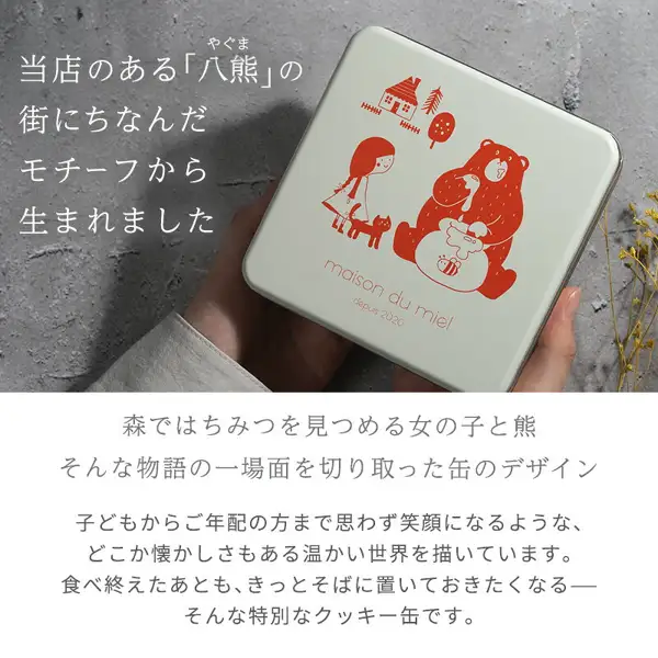 Exterior photo of the maison du miel Miel Red Cookie Tin, with a hand holding the white tin lid. The lid has red line drawings of a girl with a cat, a bear, a house and a tree, and the words “maison du miel depuis 2020”. Text the shop placed in the image reads “当店のある「八熊」の街にちなんだモチーフから生まれました” (born from motifs inspired by Yakuma, the town where our shop is located), “森ではちみつを見つめる女の子と熊 そんな物語の一場面を切り取った缶のデザイン” (a tin design capturing a scene from a story of a girl and a bear looking at honey in the forest), and “子どもからご年配の方まで思わず笑顔になるような、どこか懐かしさもある温かい世界を描いています。食べ終えたあとも、きっとそばに置いておきたくなる―そんな特別なクッキー缶です。” (it depicts a warm, somewhat nostalgic world that makes everyone from children to seniors smile; a special cookie tin you will want to keep nearby even after you finish eating)