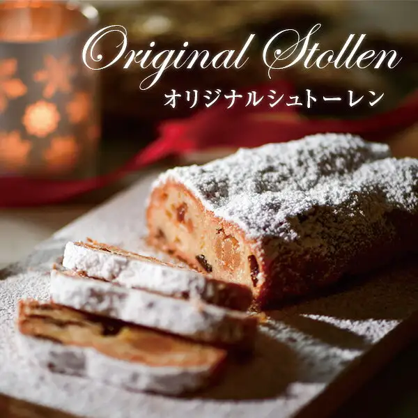 Product photo of a powdered-sugar-dusted stollen loaf with a few cut slices on a wooden board, with a red ribbon and a candle in the background (オーセントホテル小樽 Original Stollen in a Gift Box). A serving example in the cut state. Text added to the image by the shop: “Original Stollen”, “オリジナルシュトーレン”
