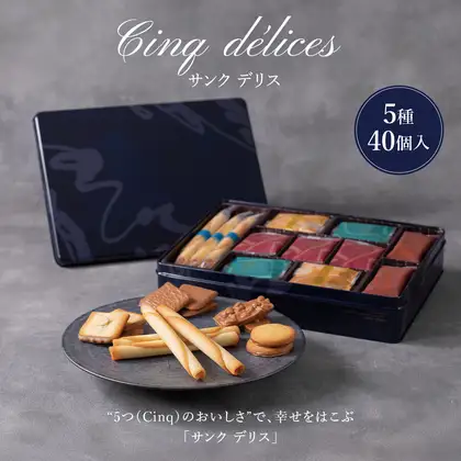 ヨックモック サンク デリス 5种40个装的商品照片。后方斜靠着藏青色的罐盖；右侧打开的罐子里分格摆放着独立包装的シガール和4种曲奇的小袋；前方灰色盘子上摆放着4根シガール和4种曲奇（ビエ オゥ ショコラオレ・ビエ オザマンド ショコラ・プラン リュンヌ・バトー ドゥ マカダミア）（摆盘示例，盘子不包含在商品内）。店铺在图片中加入的标示「Cinq délices」「サンク デリス」「5種 40個入」「“5つ(Cinq)のおいしさ”で、幸せをはこぶ」「「サンク デリス」」