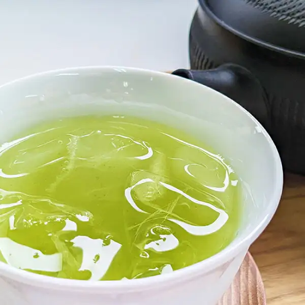 長峰製茶 さつまの風を水出しでいれたお茶の写真。白い湯のみに氷が浮かぶ黄緑色のお茶が入り、奥に黒い急須がある。