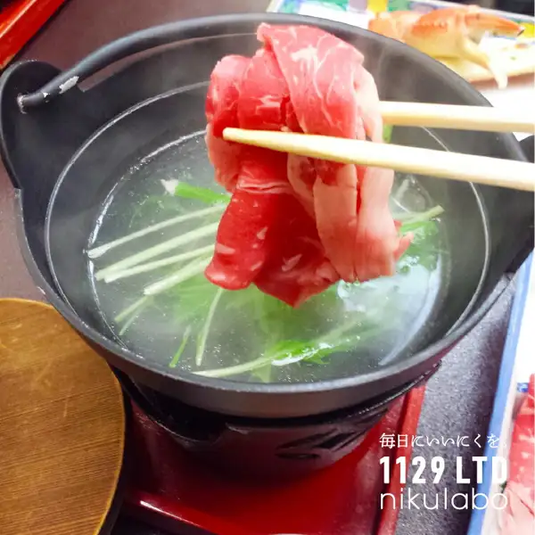 筷子夹着牛肉薄片、在放有水菜的涮涮锅汤中涮煮的烹调示例照片（黑毛和牛×かごしま黒豚 しゃぶしゃぶセット 1kg）。背景中可见疑似蟹腿的食物。店铺加在图片上的标示：“毎日にいいにくを。”（每天都吃好肉）、“1129 LTD nikulabo”