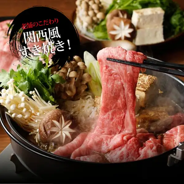 黒い鉄鍋で、箸で持ち上げた薄切りの牛肉を、えのきだけ・しめじ・しいたけ・豆腐・青菜・ねぎと一緒に煮ているすき焼きの盛り付け例の写真(うし源本店 大和榛原牛 シンタマスライス すき焼き用 800g)。店舗が画像に入れた表示「老舗のこだわり 関西風すき焼き!」。きのこ・豆腐・青菜・ねぎは商品に含まれない