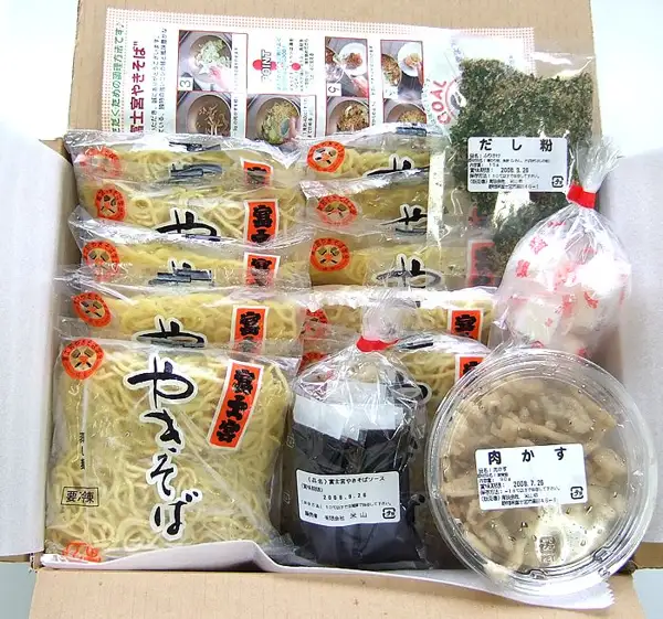 Product photo of the Tokusen Fujinomiya Yakisoba 10-Serving Set (Yoneyama): a cardboard box packed with bags of steamed noodles, a bag of dashi powder, a tub of nikukasu, a bag of sauce and a cooking-instruction sheet. The labels read “だし粉”, “肉かす” and “富士宮やきそばソース”.