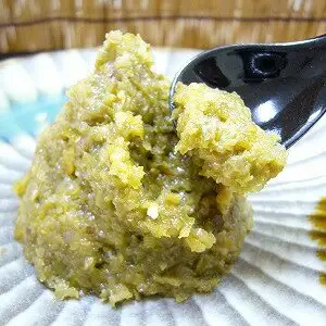 Featured image for 川津家謹製 Granular Yuzu Pepper (Green) 100g. A product photo of granular green yuzu pepper paste heaped on a white dish, with a black spoon lifting up a portion. There is no text in the image.